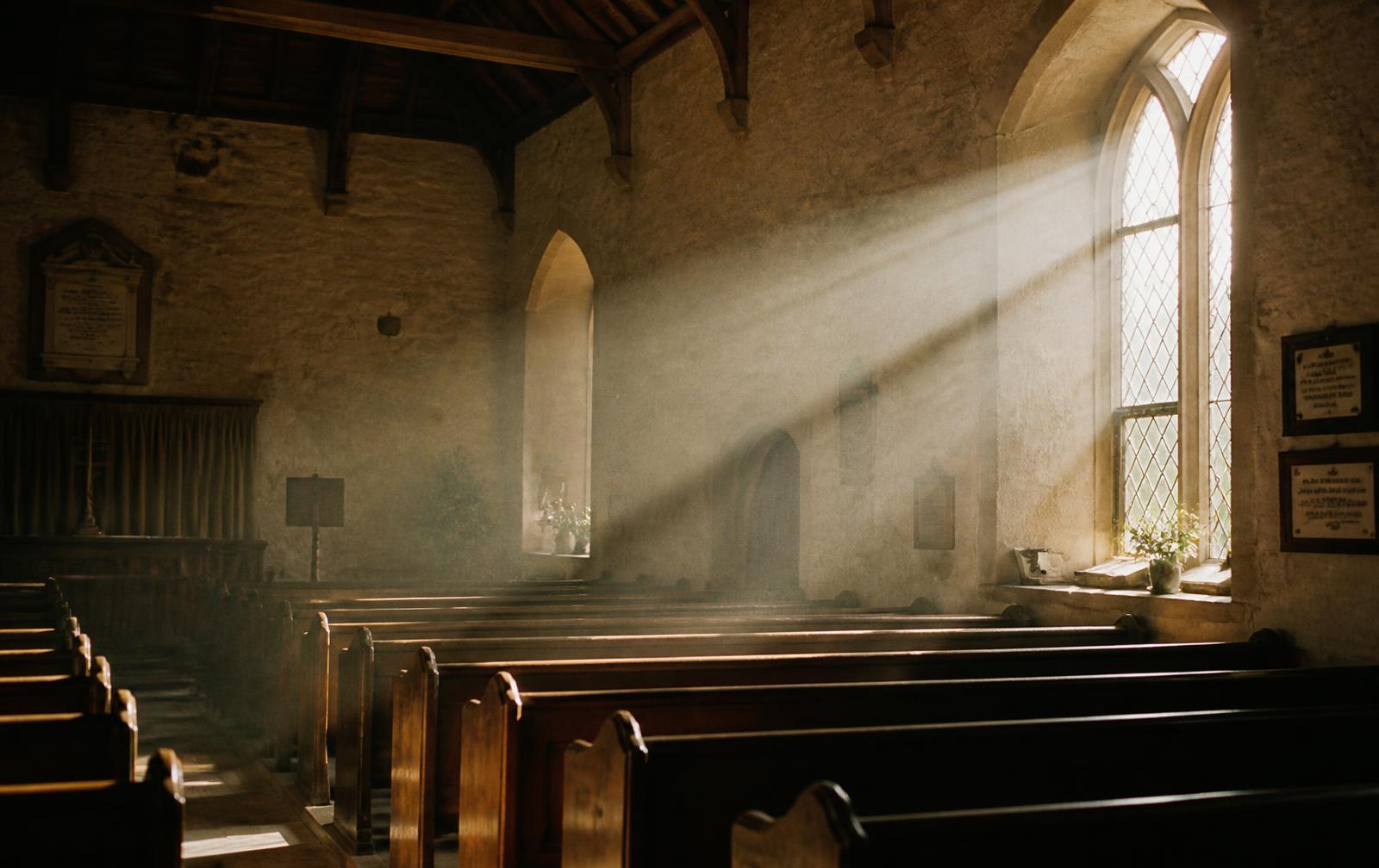 Sunlight falling through a tall arched window onto empty wooden pews in a small chapel